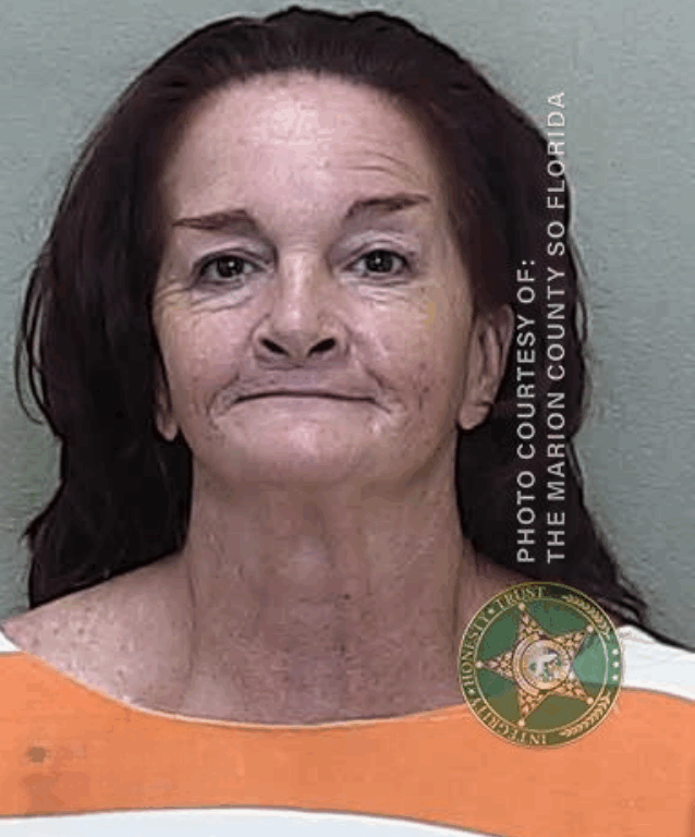 Mugshot of a middle-aged woman with long dark hair and a faint smile. She is wearing an orange and white jail uniform. The image is watermarked with “Photo Courtesy of: The Marion County SO Florida” and includes the sheriff's badge emblem.