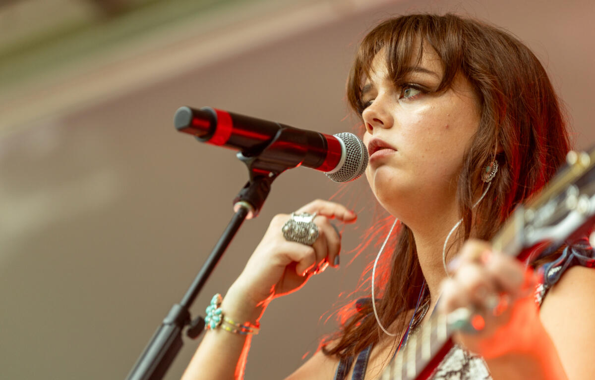 Close-up of a young woman singing into a microphone while playing guitar on stage.