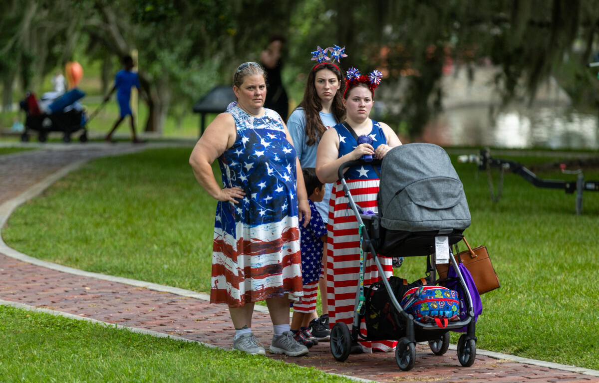 Three women in American flag-themed clothing stand with a stroller and child during a park celebration.