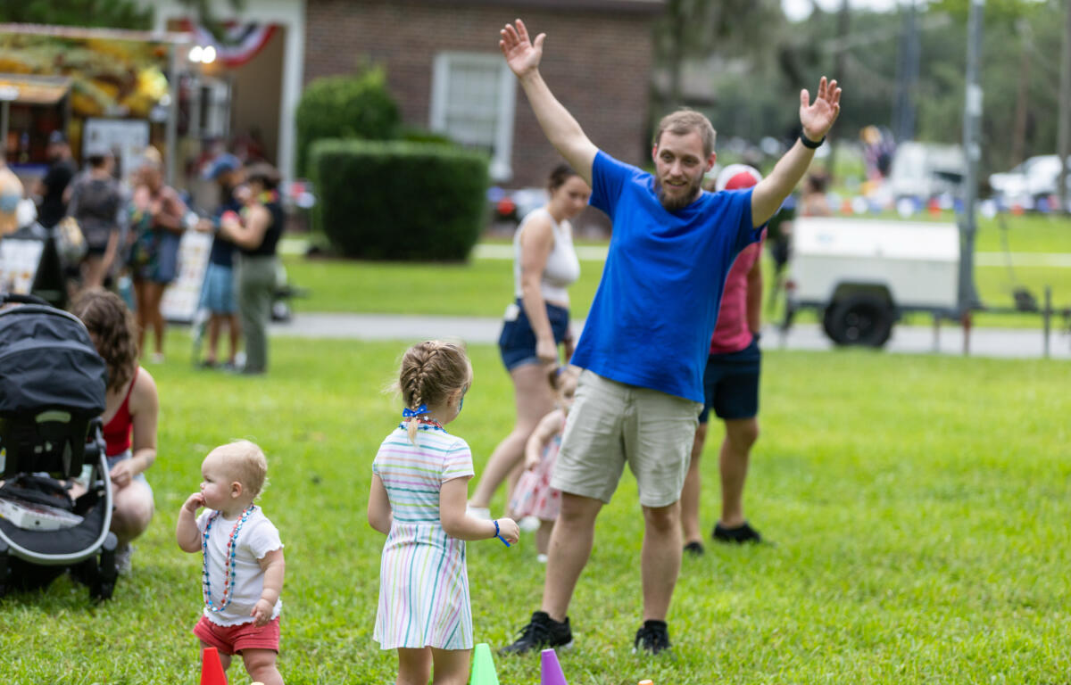Man raises arms as young children wearing patriotic necklaces play near colorful cones on the grass during a public event.