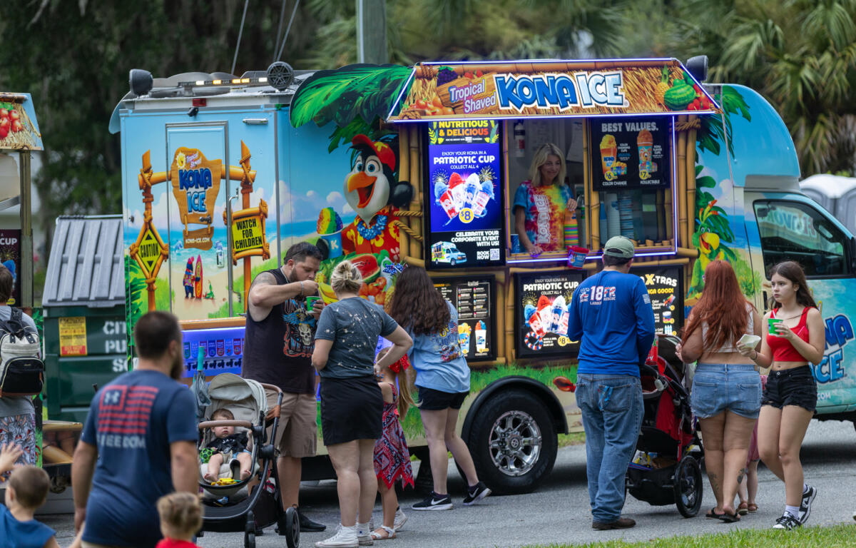 People gather around a colorful Kona Ice truck, ordering shaved ice during a community celebration.