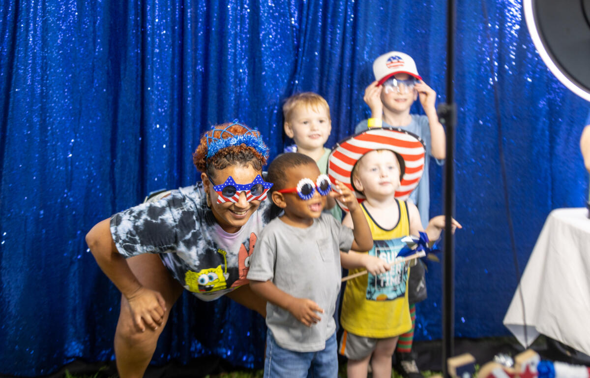 Smiling children and adult wear red, white, and blue sunglasses and hats in front of a sparkly blue backdrop.