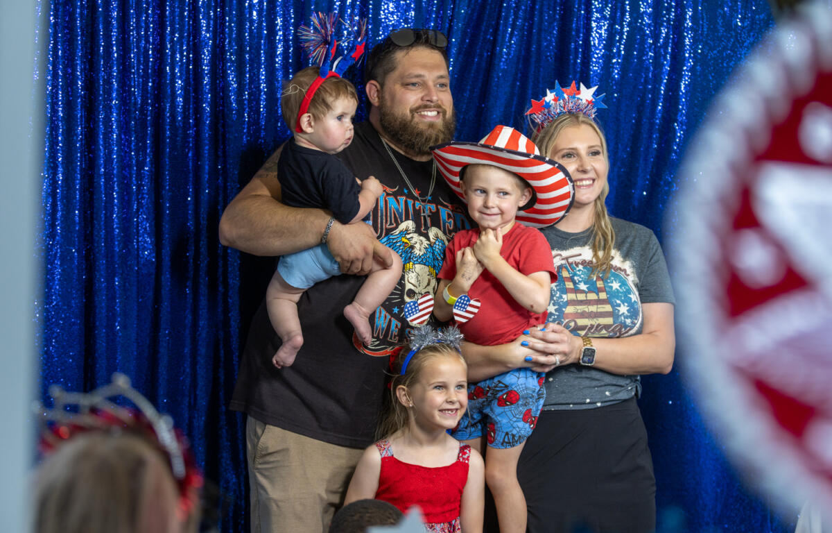 Smiling family of five dressed in red, white, and blue outfits pose in front of a blue sequin backdrop with festive accessories.