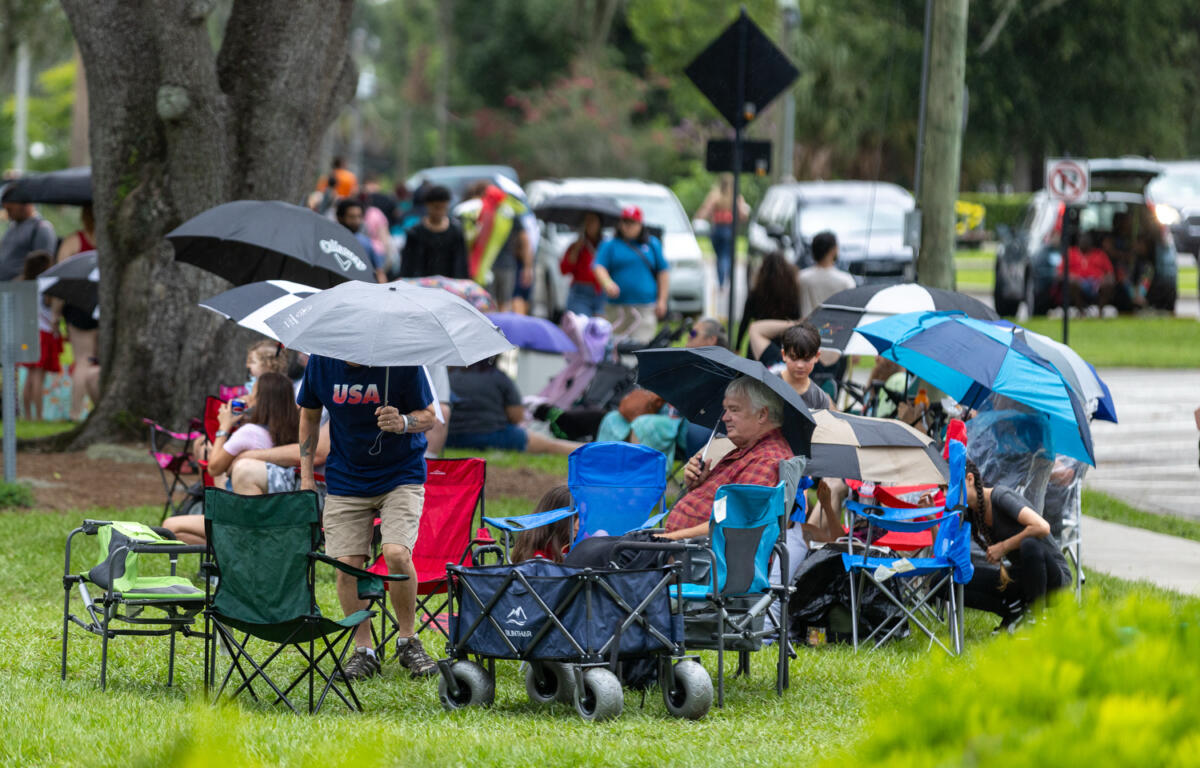 People with umbrellas and folding chairs gather at Tuscawilla Park in Ocala, Florida, during rainy Fourth of July festivities.
