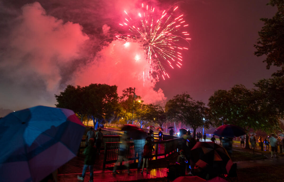 Crowd watches red fireworks burst in the rainy night sky with umbrellas open at a park celebration.