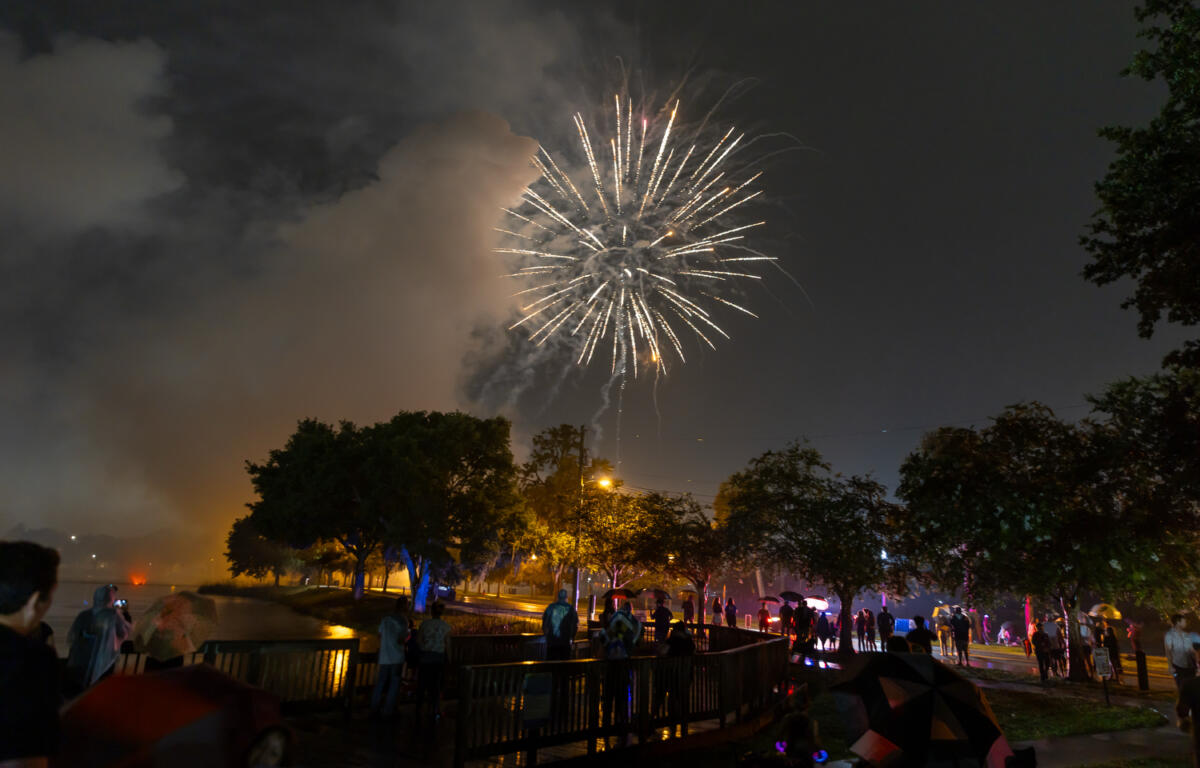 People with umbrellas watch golden fireworks light up the rainy night sky at a park celebration.