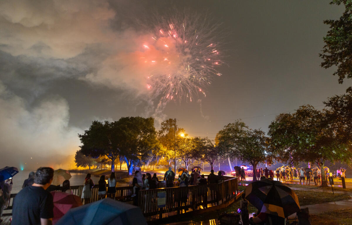 People holding umbrellas watch fireworks explode in a cloudy sky during a Fourth of July celebration at a park.