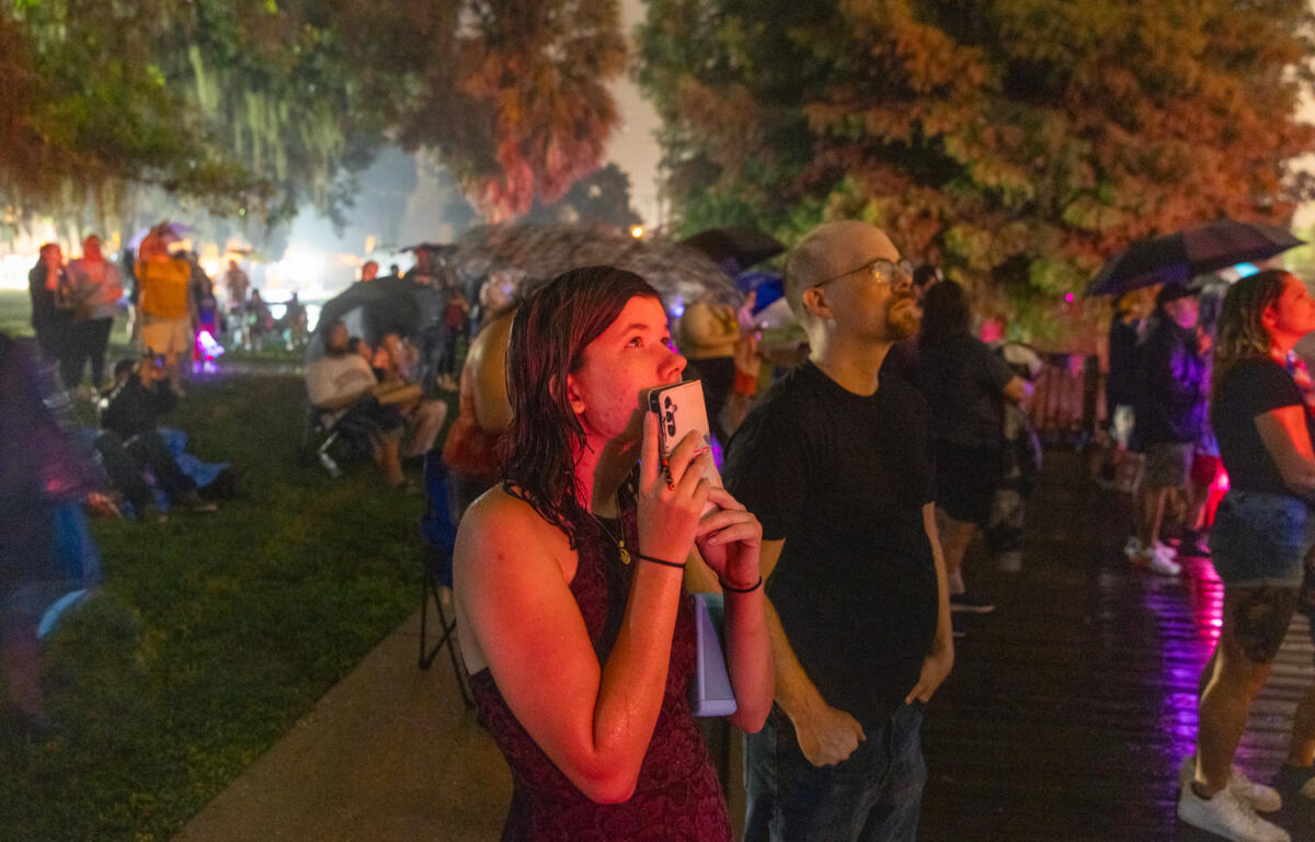 Wet woman holding phone smiles while watching fireworks during a rainy Fourth of July celebration.