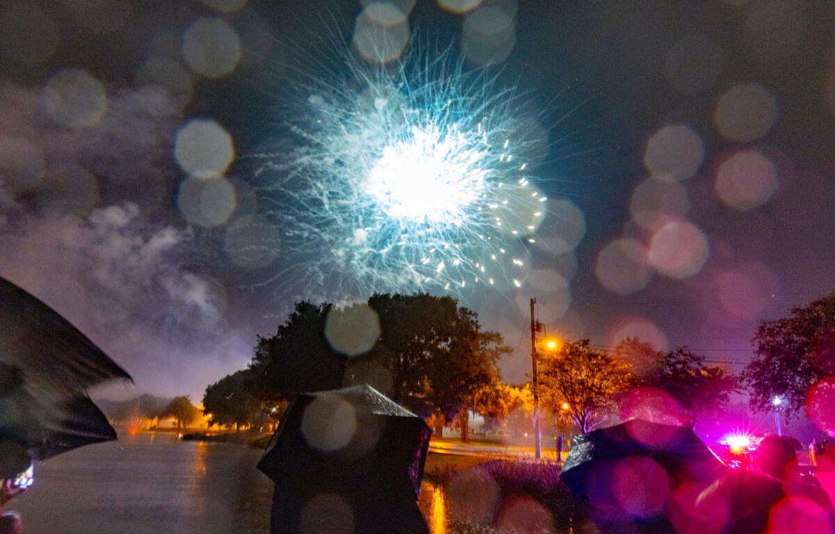 Umbrellas and raindrops fill the frame as a bright blue firework lights up the rainy night sky at a park celebration.