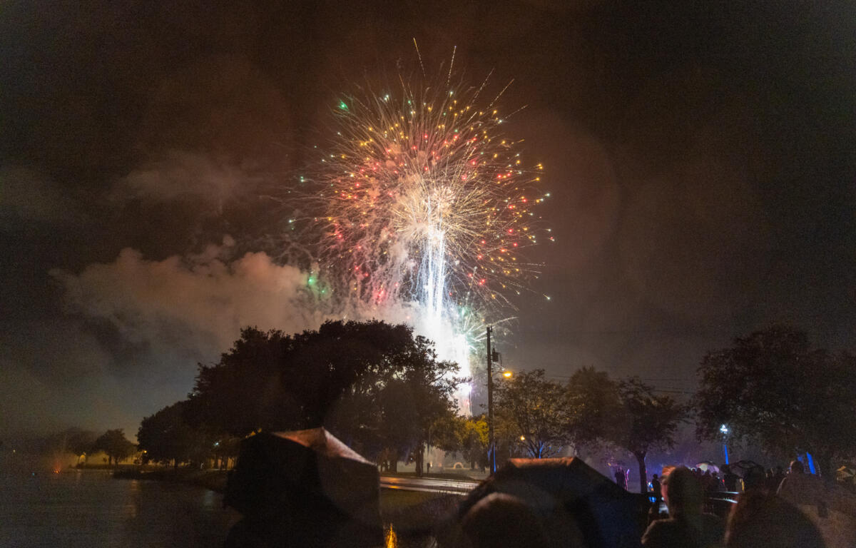 Large multicolored fireworks explode in the night sky above trees and umbrellas during a holiday celebration.