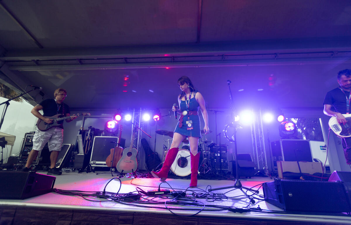 Female vocalist in red boots stands onstage with microphone as band members play guitar and bass under colorful stage lights.