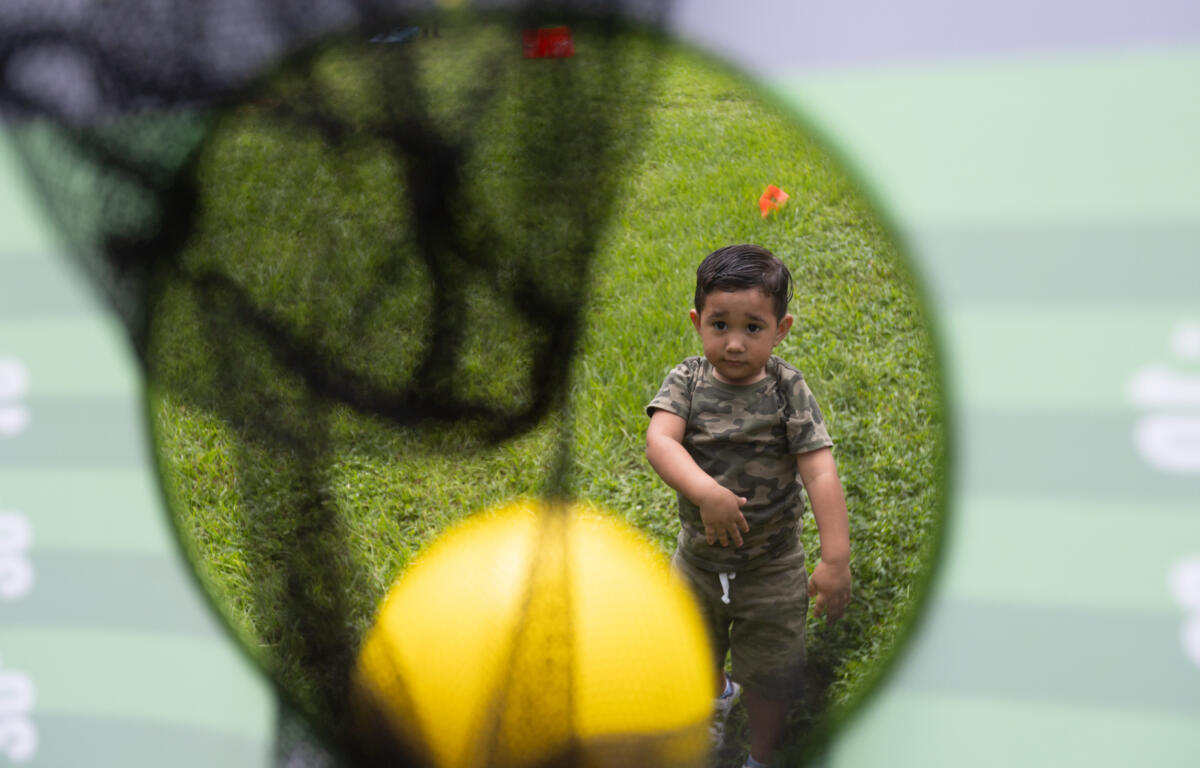 Toddler in camouflage shirt watches a yellow ball fall into a net during an outdoor game on grass.