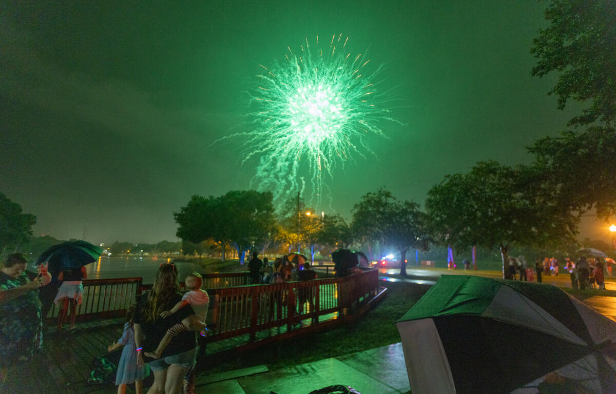 People with umbrellas watch green fireworks in the sky over a wet park during a nighttime Fourth of July celebration.