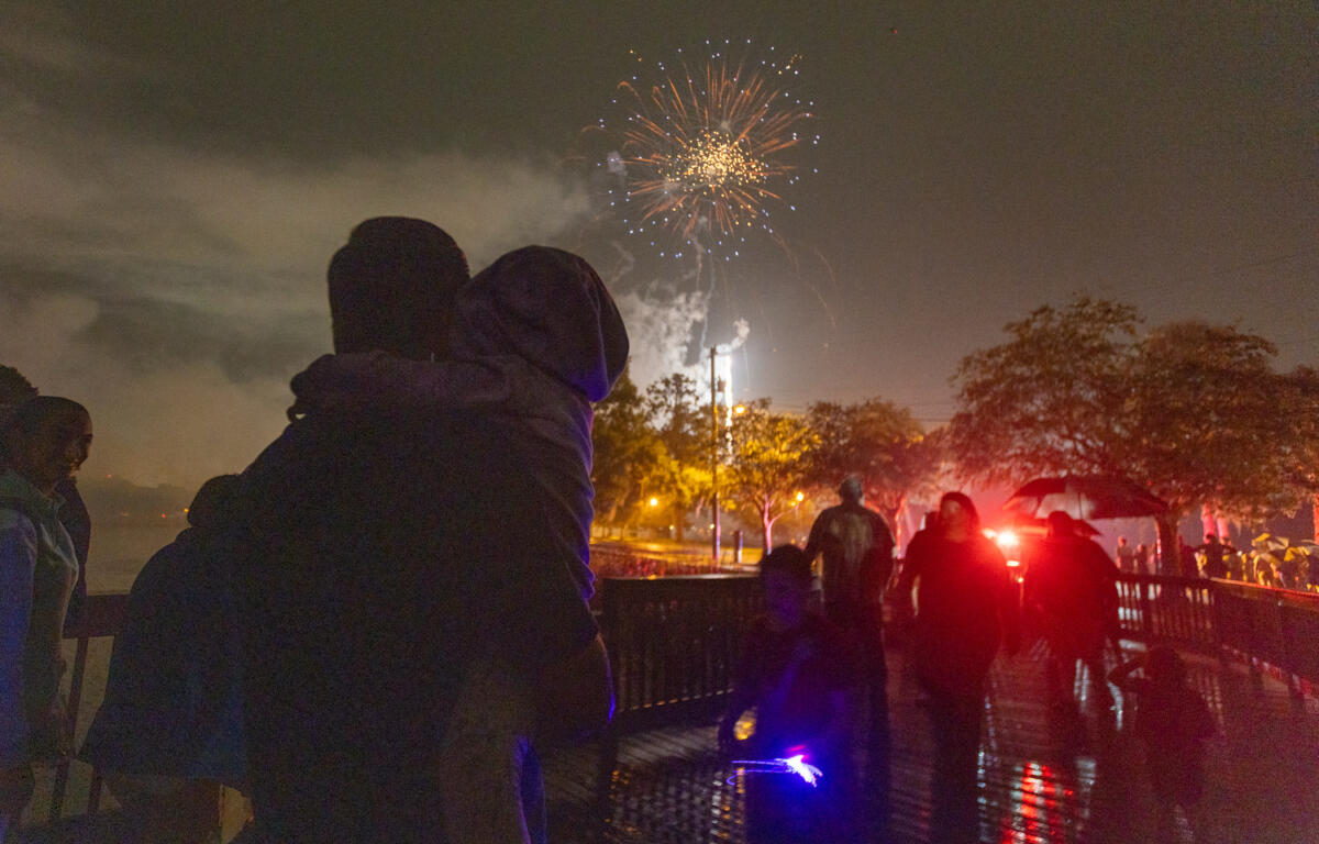 Silhouetted family and crowd watch fireworks explode in the sky on a rainy night in Ocala during a Fourth of July event.