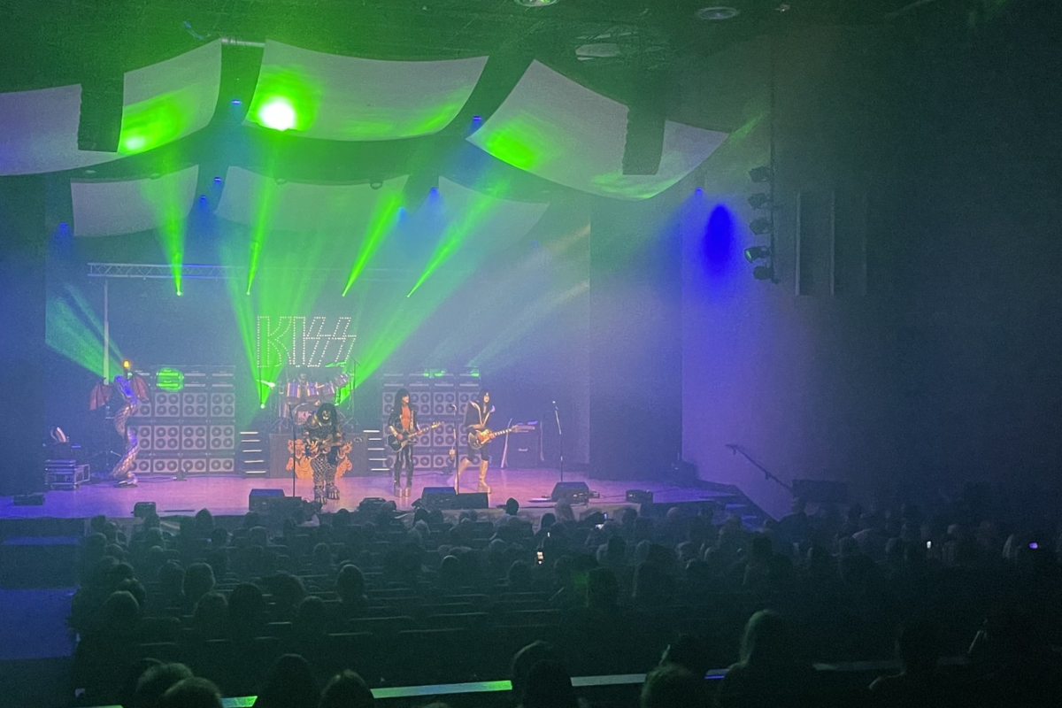 A rock tribute band dressed in full KISS costumes performs on stage under green and blue lights at a concert venue, with the illuminated KISS logo behind them and a seated audience watching the show.