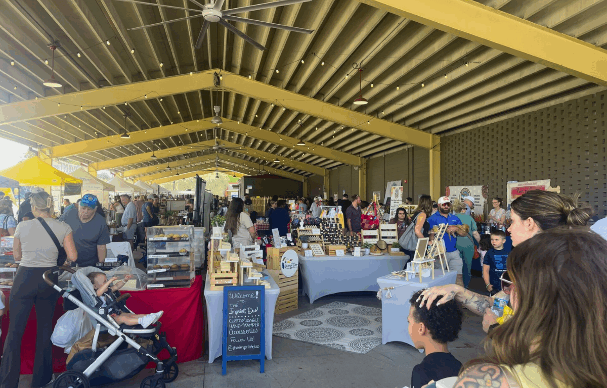 Shoppers browse vendor booths under a large yellow pavilion at the Ocala Downtown Market, featuring handmade goods, baked treats, and local produce on display.