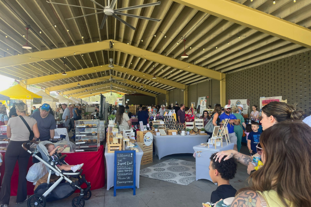 Shoppers browse vendor booths under a large yellow pavilion at the Ocala Downtown Market, featuring handmade goods, baked treats, and local produce on display.