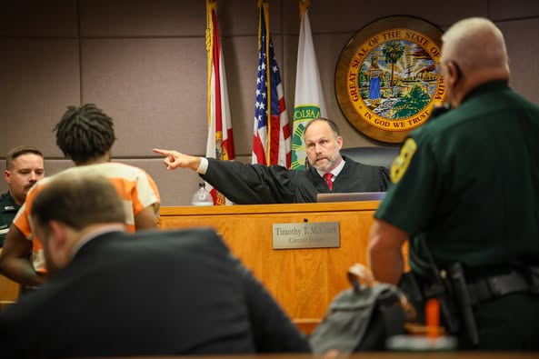 Judge points during courtroom sentencing as defendant in orange jumpsuit stands before the bench, surrounded by law enforcement and legal personnel.
