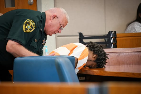 A sheriff's deputy leans over a courtroom table as the defendant, in a striped uniform, rests his head face-down on the surface.