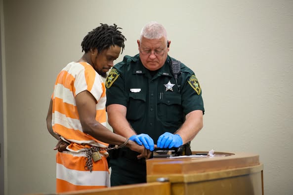 A sheriff's deputy holds the hand of the defendant, getting his fingerprints recorded.