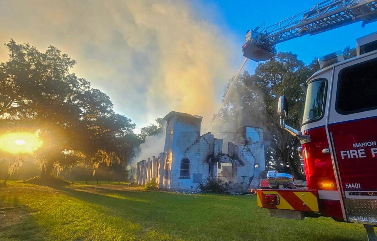Firetruck and ladder spraying water onto smoldering remains of a burned church as morning sun rises behind nearby trees.