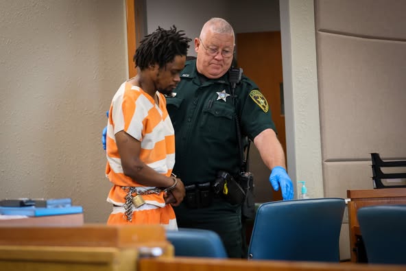 A man in an orange and white striped jail uniform stands in a courtroom with his hands cuffed in front of him, accompanied by a Marion County Sheriff's deputy wearing gloves.