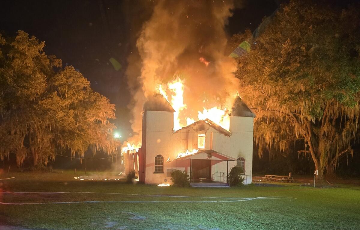 Front view of church engulfed in flames with fire shooting from the roof and smoke rising into the night sky.
