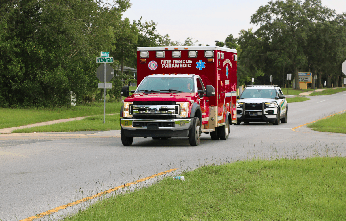 A red truck with "Fire Rescue Paramedic" written on the front and a white cop car driving down a road surrounded by trees at a stop sign.