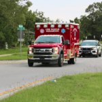 A red truck with "Fire Rescue Paramedic" written on the front and a white cop car driving down a road surrounded by trees at a stop sign.
