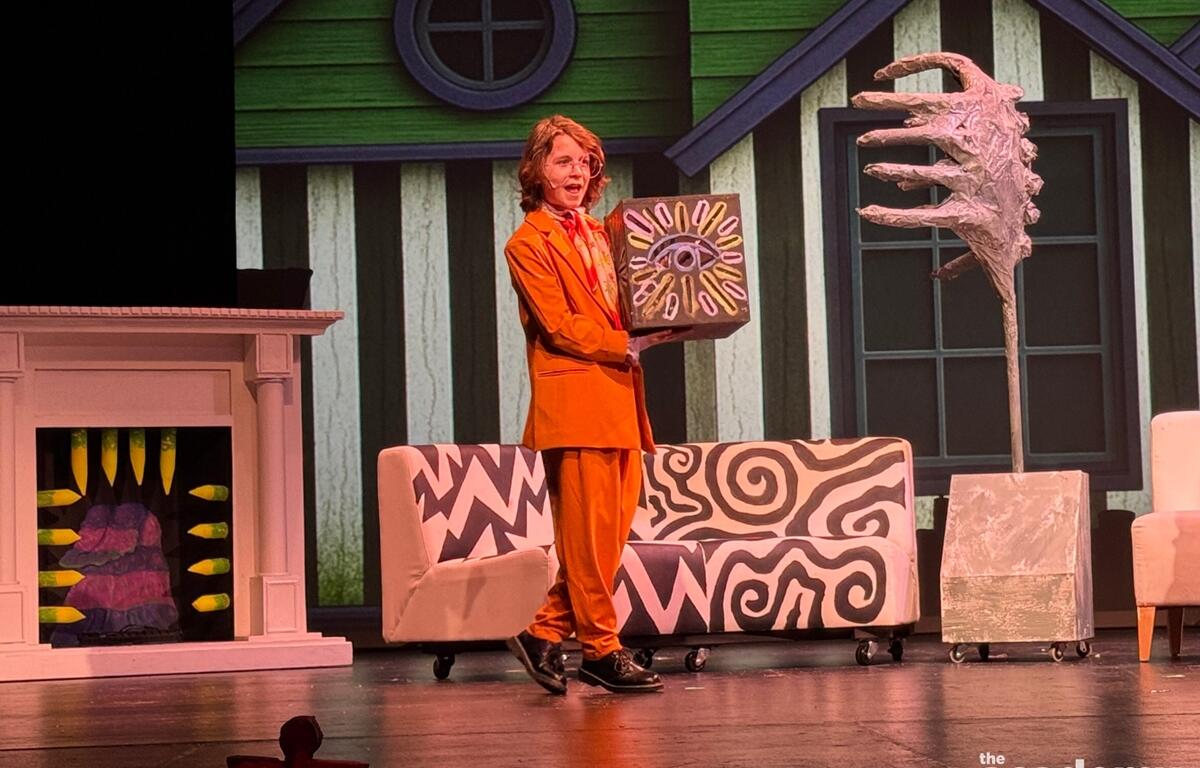 Youth actor in orange suit performs on stage during Beetlejuice Jr. in front of a colorful set.