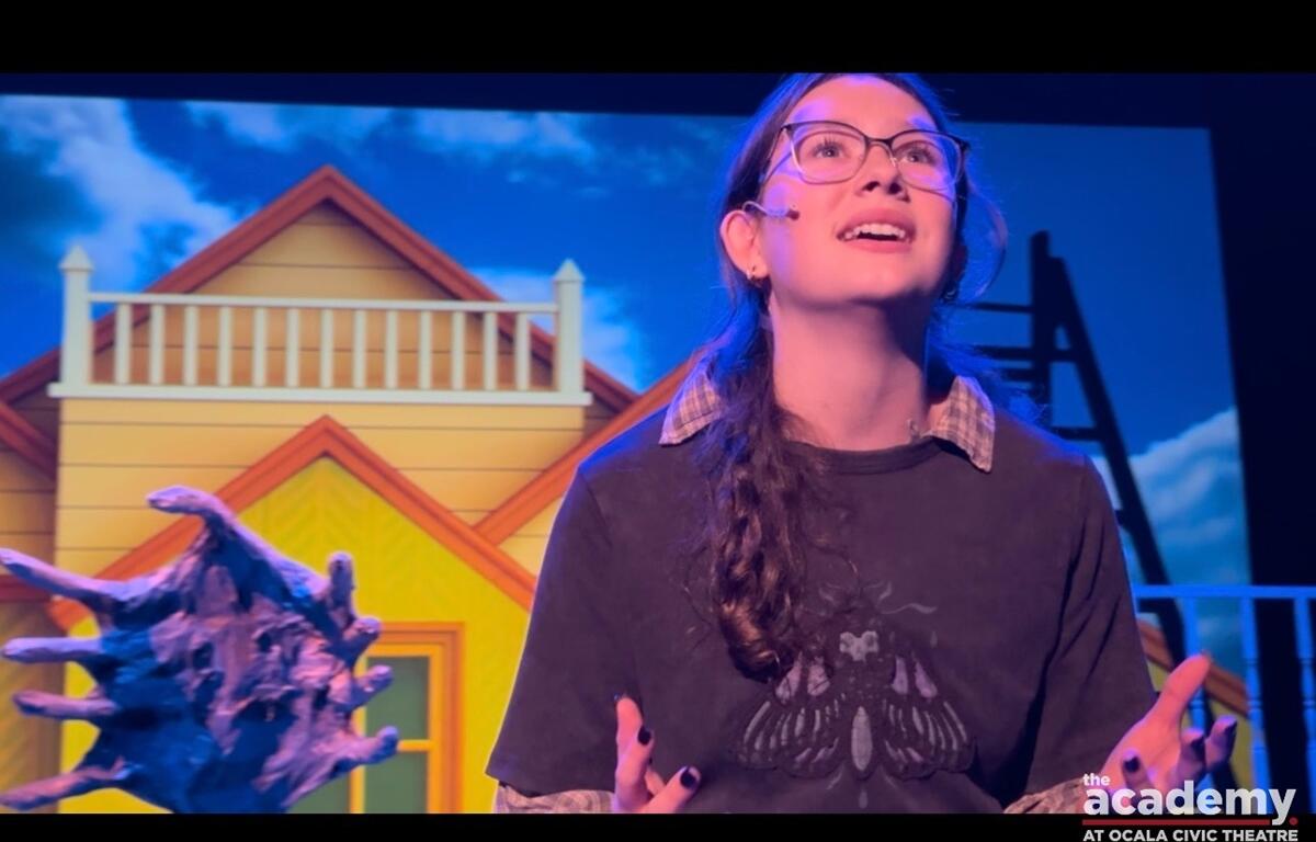 Youth actor portraying Lydia performs on stage in front of a colorful house backdrop during Beetlejuice Jr.