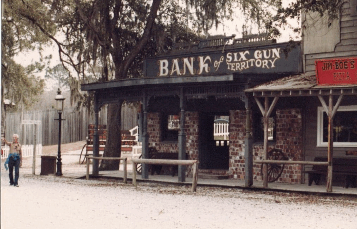 A brick building donning a sign that reads, "Bank of Six Gun Territory" is housed on a street corner next to Jim-Boe's Pizza. 