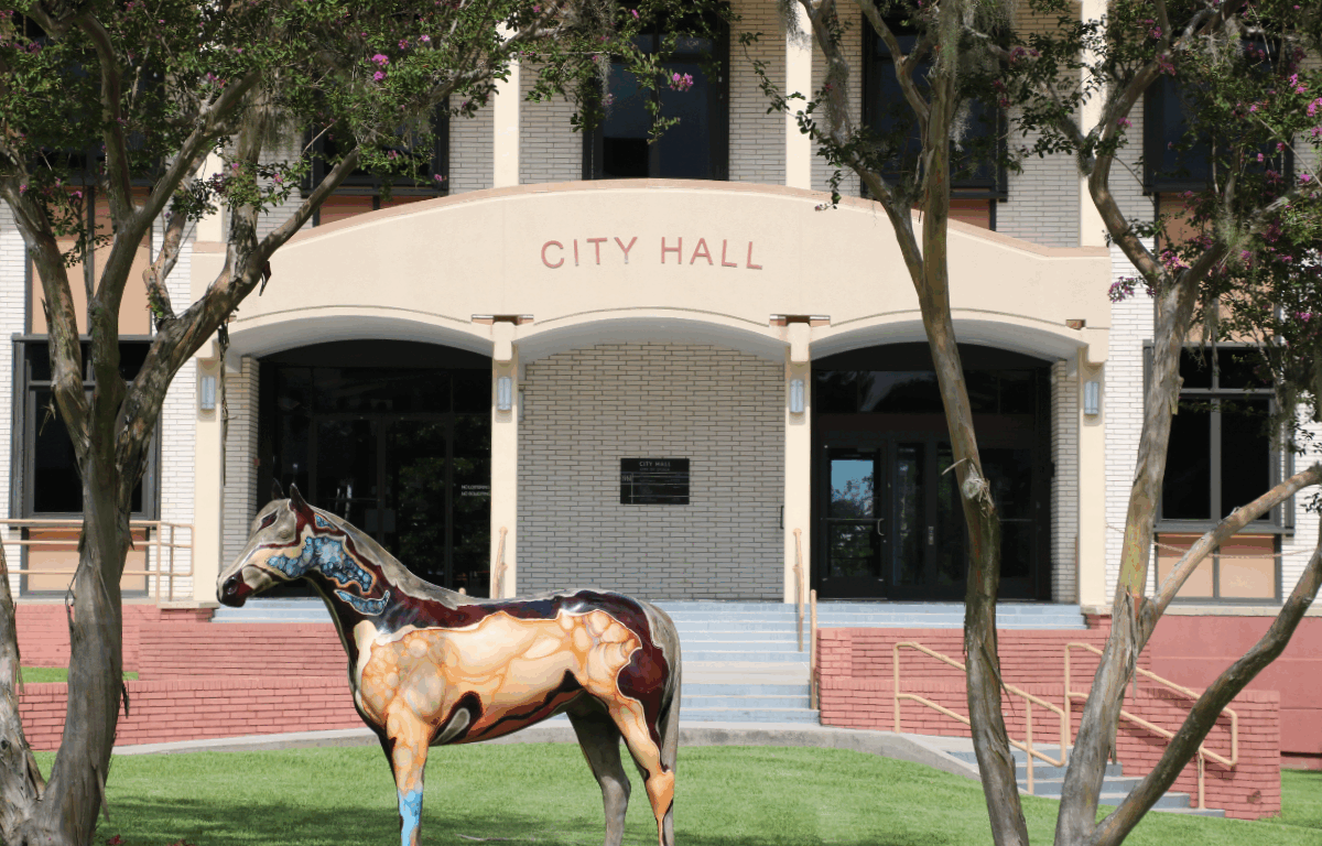 The front entrance of Ocala’s City Hall in the background with “CITY HALL” displayed on an arch above the double door entrances on the left and right. There are short red brick walls in front of the entrance with stairs leading up to the doors on either side. In the foreground are trees on either side of the City Hall arch and a beige and blue horse statue facing left.