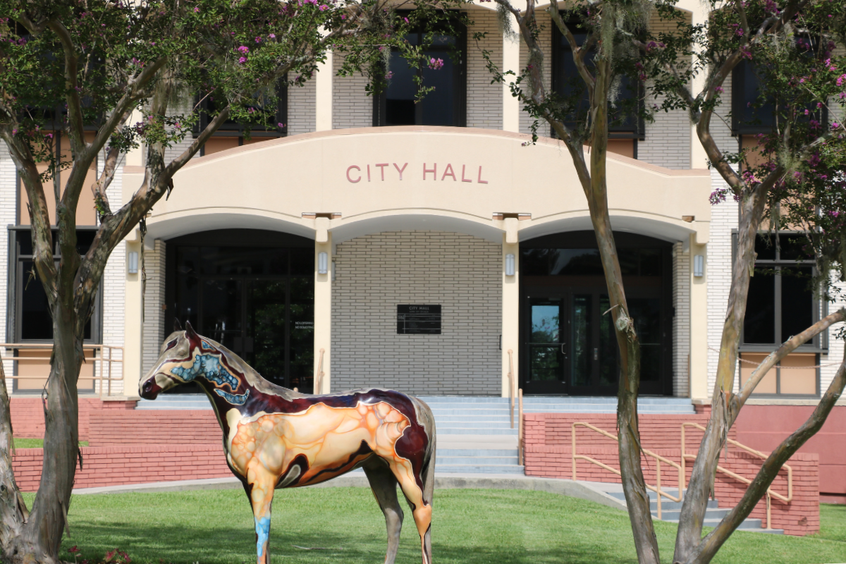 The front entrance of Ocala’s City Hall in the background with “CITY HALL” displayed on an arch above the double door entrances on the left and right. There are short red brick walls in front of the entrance with stairs leading up to the doors on either side. In the foreground are trees on either side of the City Hall arch and a beige and blue horse statue facing left.