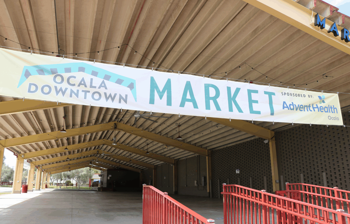 A large banner reading “Ocala Downtown Market" hangs under the roof of a covered pavilion. The structure features yellow steel beams and an open concrete floor, with string lights hanging from the ceiling. Red railings line the entrance, and trees and grass are visible in the background.