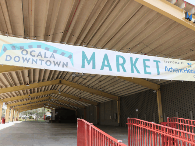 A large banner reading “Ocala Downtown Market" hangs under the roof of a covered pavilion. The structure features yellow steel beams and an open concrete floor, with string lights hanging from the ceiling. Red railings line the entrance, and trees and grass are visible in the background.