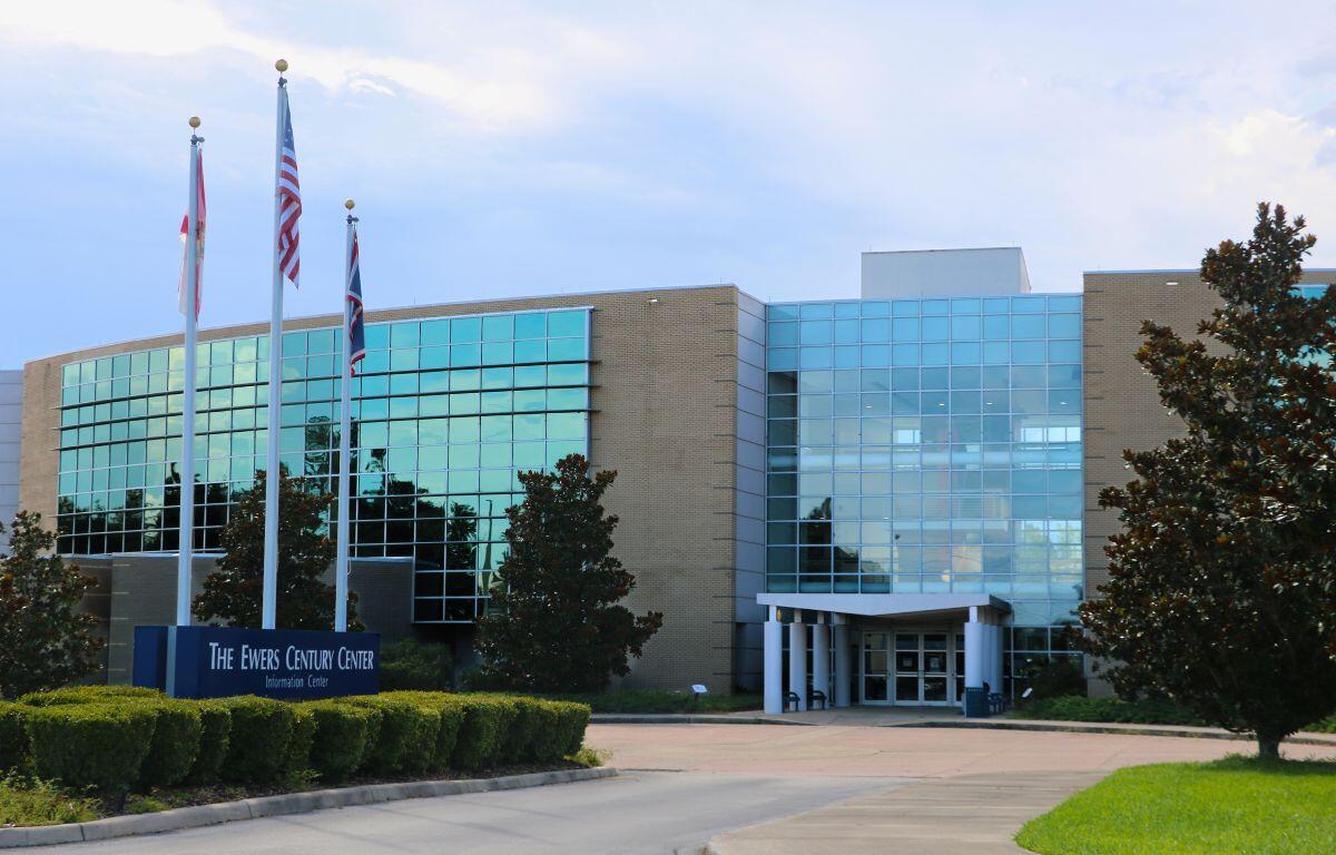 The entrance facade of The Ewers Century Center, a modern glass-and-brick building with large reflective windows. In front are trimmed bushes, magnolia trees, and a banner reading “The Ewers Century Center Information Center.”