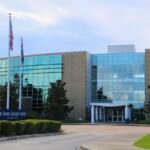 The entrance facade of The Ewers Century Center, a modern glass-and-brick building with large reflective windows. In front are trimmed bushes, magnolia trees, and a banner reading “The Ewers Century Center Information Center.”