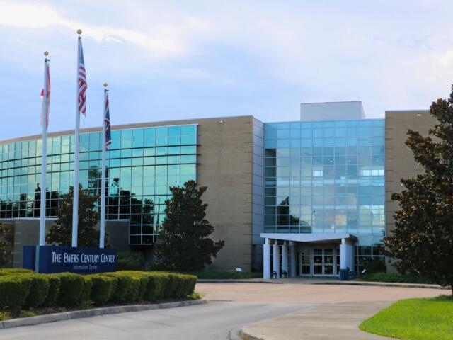 The entrance facade of The Ewers Century Center, a modern glass-and-brick building with large reflective windows. In front are trimmed bushes, magnolia trees, and a banner reading “The Ewers Century Center Information Center.”