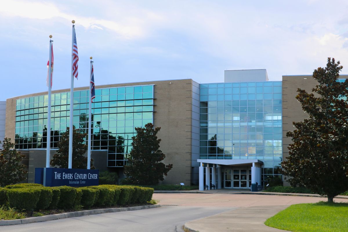 The entrance facade of The Ewers Century Center, a modern glass-and-brick building with large reflective windows. In front are trimmed bushes, magnolia trees, and a banner reading “The Ewers Century Center Information Center.”