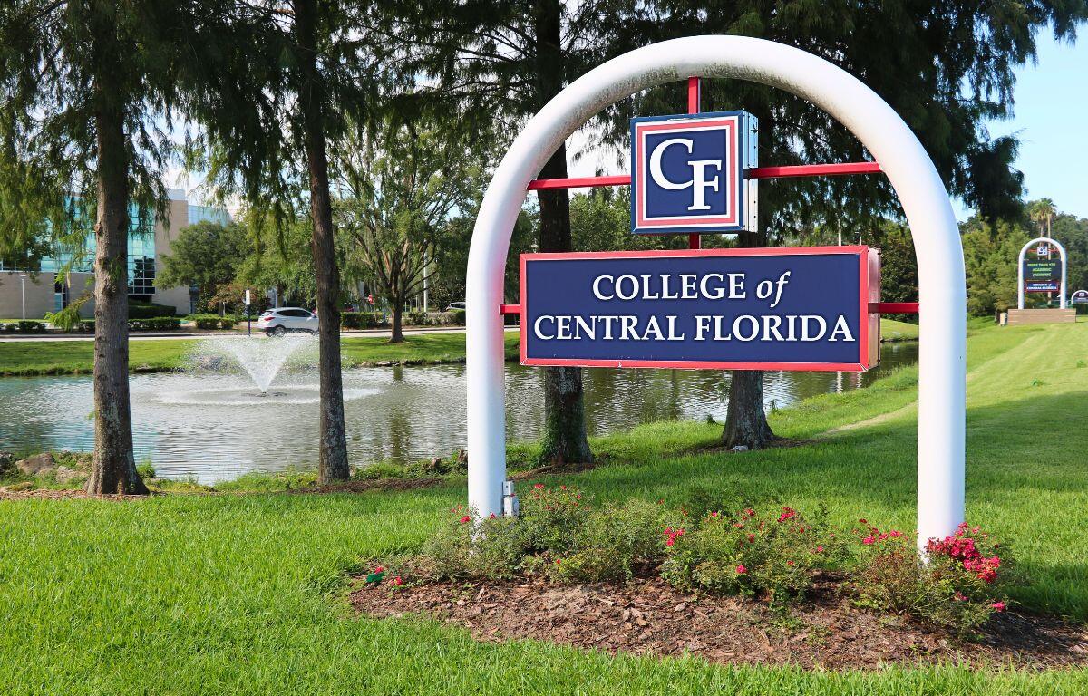 A standalone entrance sign with a white arch and red structural supports, displaying a blue panel with “CF” and below it “College of Central Florida.” Behind it is a small pond with a fountain and a grassy lawn.