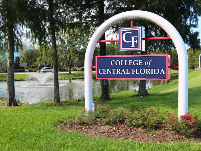 A standalone entrance sign with a white arch and red structural supports, displaying a blue panel with “CF” and below it “College of Central Florida.” Behind it is a small pond with a fountain and a grassy lawn.