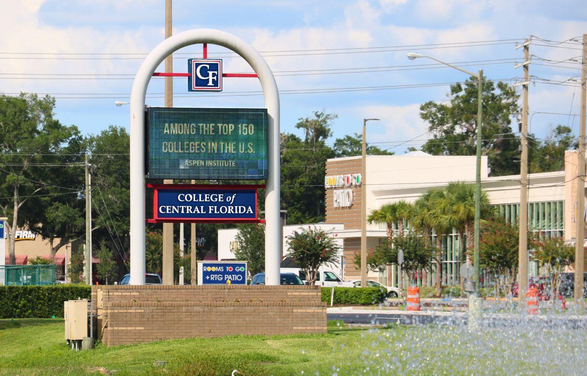A wide shot showing a College of Central Florida sign with an LED display in front of a landscaped area. In the distance are a pond fountain, a large Rooms To Go Patio building, trees and traffic cones.
