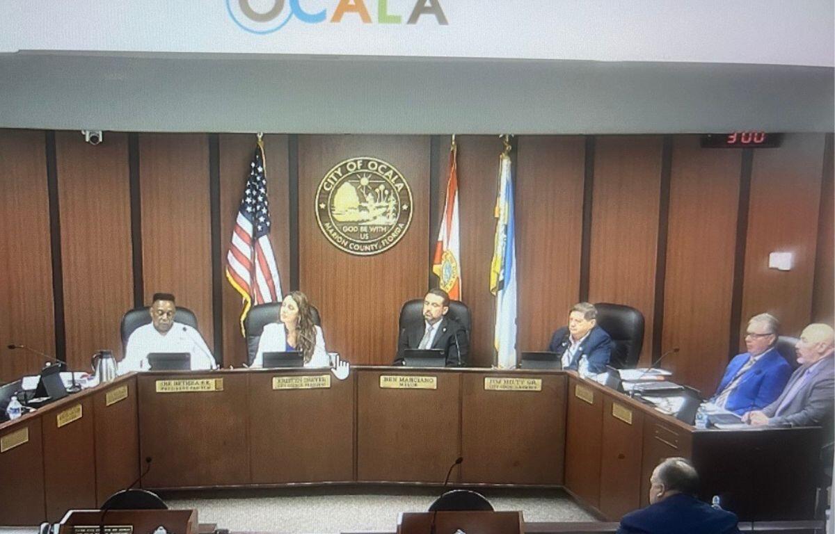 A group pf well-dressed people sitting behind a raised dais platform, with flags to either side of an insignia with letter, numbers and an image, and a city of Ocala logo against a white background.