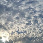 A bright blue sky covered in scattered clouds with sunlight shining faintly through them.