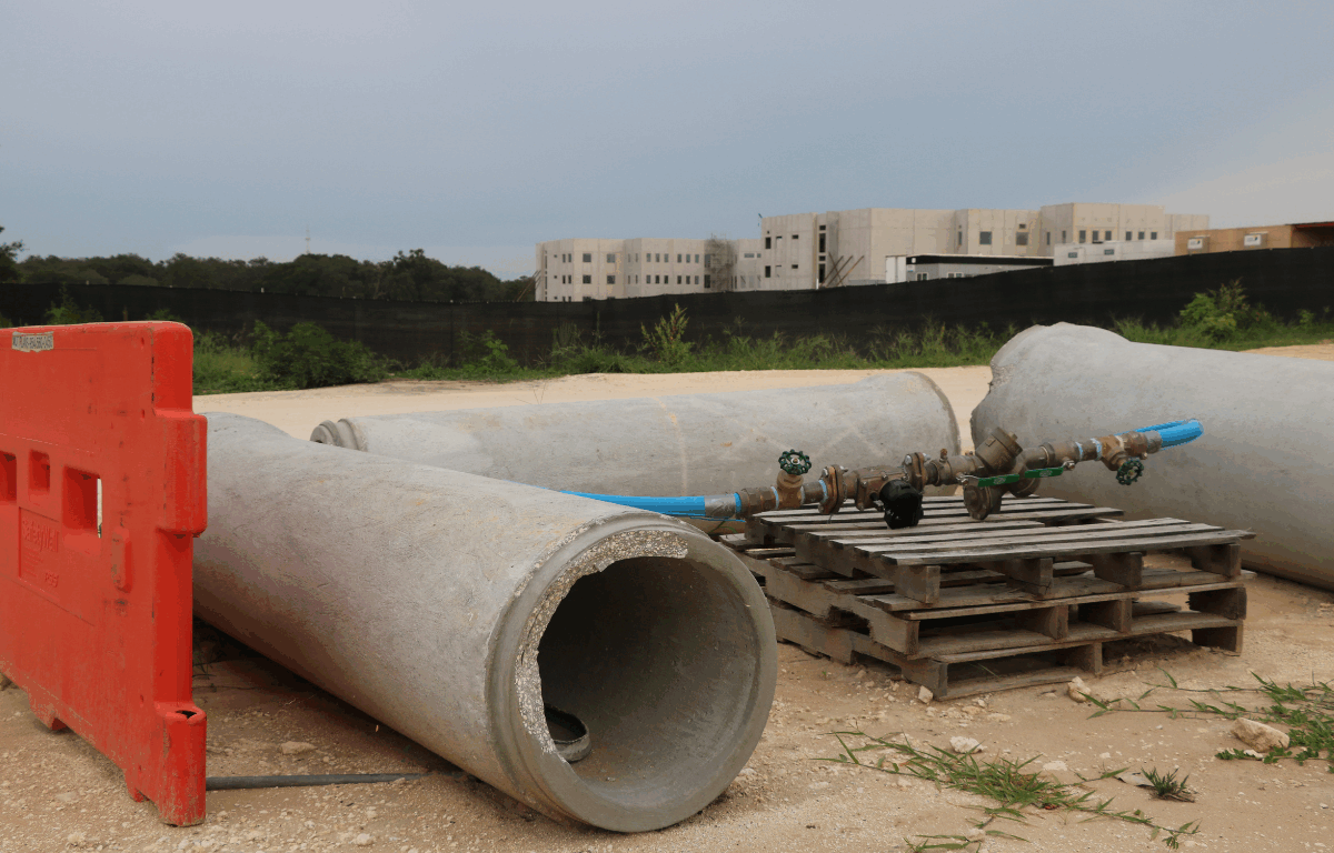 In the background, a blurred view of a building under construction stands behind a tall fence with a black tarp in front of it and trees to the left of it. In the foreground are various construction tools including wood pallets, concrete tubes, an orange barrier and a water valve.