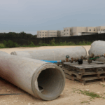 In the background, a blurred view of a building under construction stands behind a tall fence with a black tarp in front of it and trees to the left of it. In the foreground are various construction tools including wood pallets, concrete tubes, an orange barrier and a water valve.