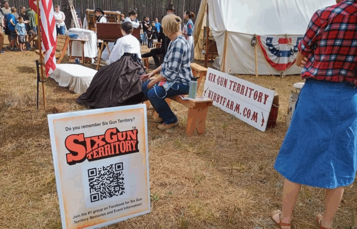 A reenactment scene at a Six Gun Territory event shows people in pioneer-era costumes gathered near a canvas tent and wooden benches. A QR code sign invites attendees to join a Facebook group, and a wooden sign directs visitors to “KirbyFarm.com.” An American flag stands nearby, and families watch the activity from the background.