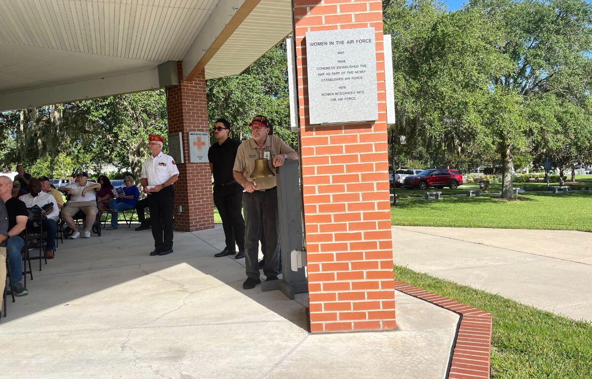 A man dressed in a uniform, stands by a bell, with people to his left and brick column with a granite plaque affixed to it to his right.