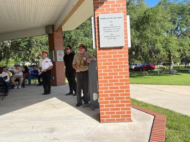 A man dressed in a uniform, stands by a bell, with people to his left and brick column with a granite plaque affixed to it to his right.